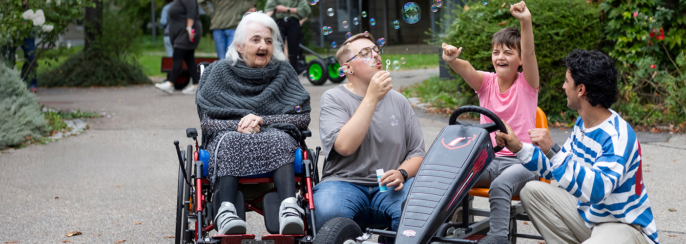 Vier Personen in unterschiedlichem Alter befinden sich nebeneinander. Davon sitzt eine Person im Rollstuhl, eine Person bläst Seifenblasen und eine weitere Person sitzt auf einem Kettcar. Vier Personen in unterschiedlichem Alter befinden sich nebeneinander. Davon sitzt eine Person im Rollstuhl, eine Person bläst Seifenblasen und eine weitere Person sitzt auf einem Kettcar.